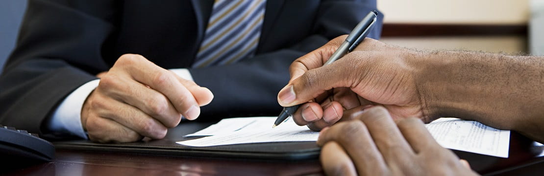 A close-up shot of the hands of two people seated across from one another a desk. One individual holds a pen above a business document.