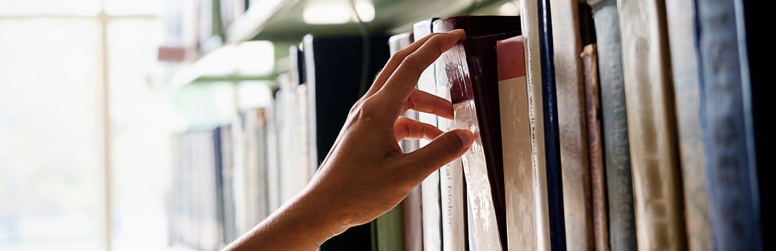 A hand takes one book off of a library shelf.